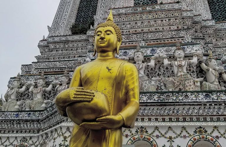 Photographie d'une statue élancée de Bouddha maigre au Wat Arun, le "Temple de l'Aube", situé à Bangkok, en Thaïlande. Le temple est connu pour ses tours majestueuses et ses mosaïques colorées.